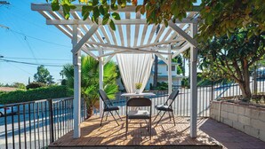 Eine überdachte Terrasse mit Tisch und Stühlen, eine weiße Pergola und Blick auf eine Straße und Grünflächen.