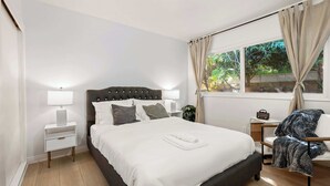 Ein ordentlich eingerichtetes Schlafzimmer mit einem großen Bett, Nachttischen, einem Sessel und einem Fenster mit Vorhängen.