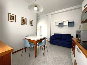 Ett kompakt vardagsrum med ett matbord och stolar, en blå soffa, en liten köksdel med mikrovågsugn och inramade tavlor på väggen.