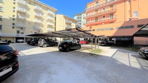 Un espace de stationnement avec plusieurs voitures, un abri-voiture couvert et des immeubles à plusieurs étages dotés de balcons.