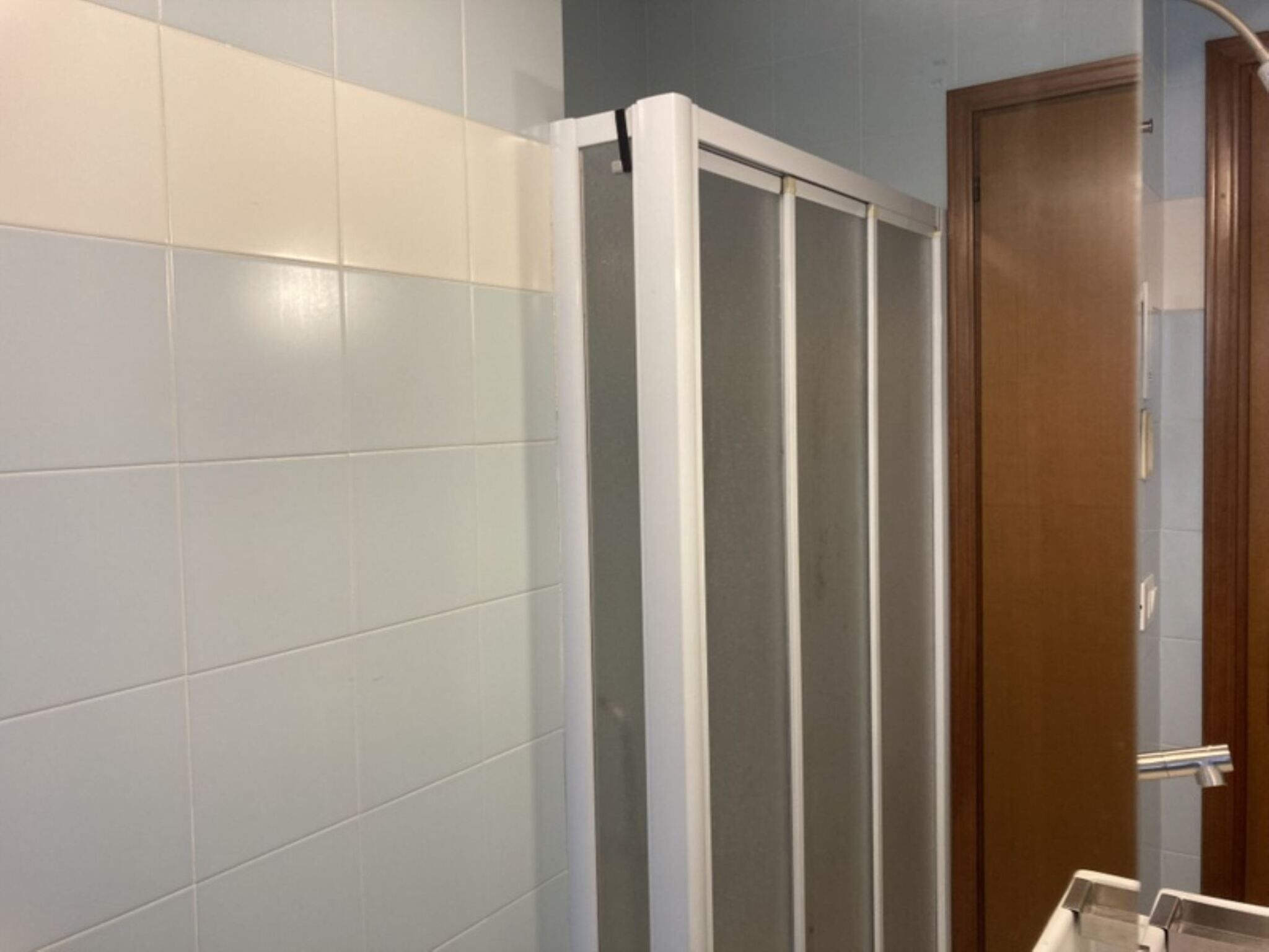 A bathroom with white and light blue tiles, a wooden door, and a shower enclosure.