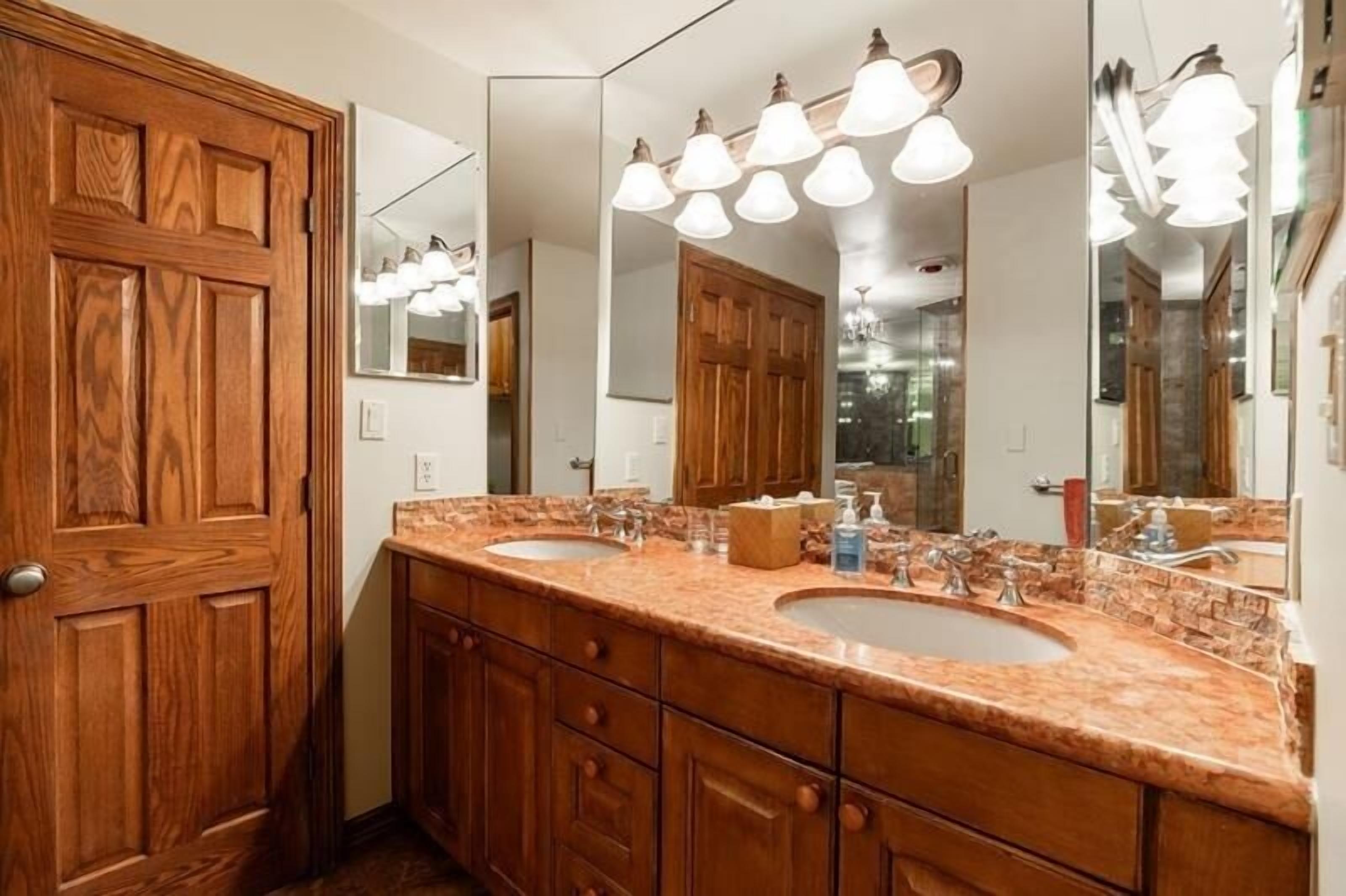 Une salle de bain avec une porte en bois, deux lavabos, un grand miroir et plusieurs luminaires.