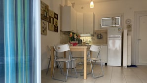 Eine kompakte Küche mit weißen Schränken, einem kleinen Esstisch mit Stühlen, einem Kühlschrank und einem Fenster mit Jalousien.