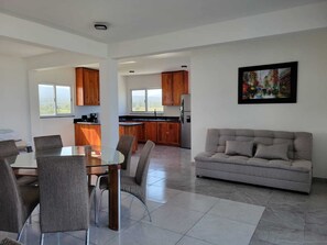 Ein modernes Wohnzimmer mit einer grauen Couch, einem Esstisch mit Stühlen und einer Küche im Hintergrund.