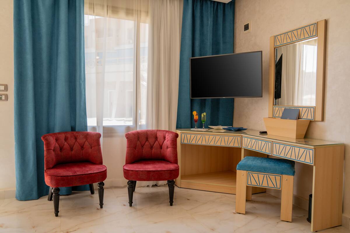 Une pièce avec un téléviseur, deux fauteuils rouges, un bureau en bois et un miroir.