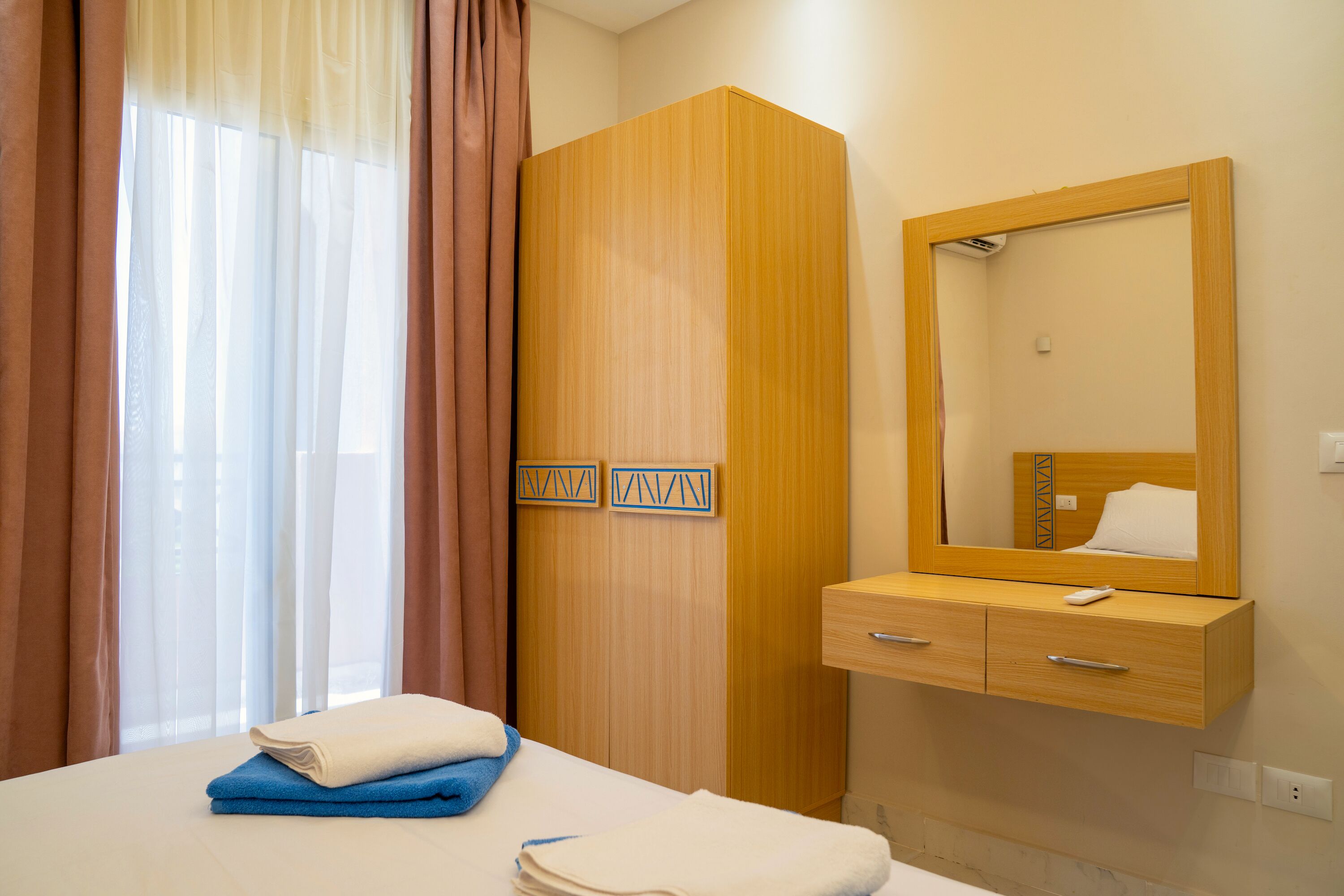 A hotel room with a bed, a wooden wardrobe, a mirror, and a window with curtains.