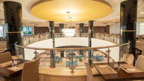 Un amplio vestíbulo de hotel con una zona de estar circular central, varias mesas con sillas y un diseño de techo moderno.