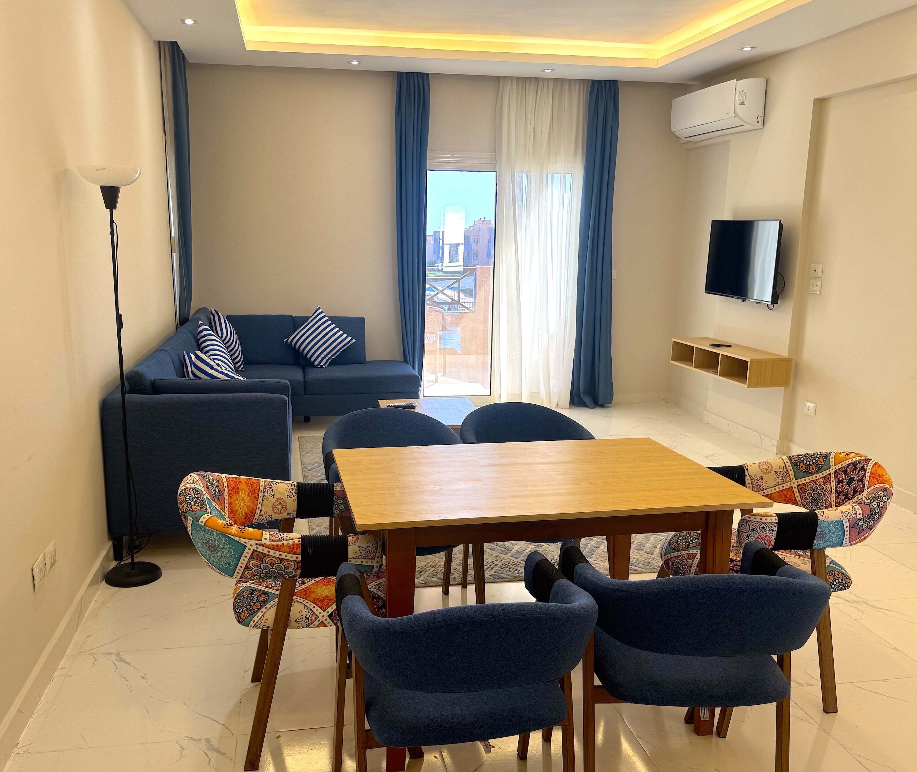 Uma sala de estar moderna com sofá, mesa de jantar e cadeiras. Há uma TV fixada na parede e um aparelho de ar condicionado.