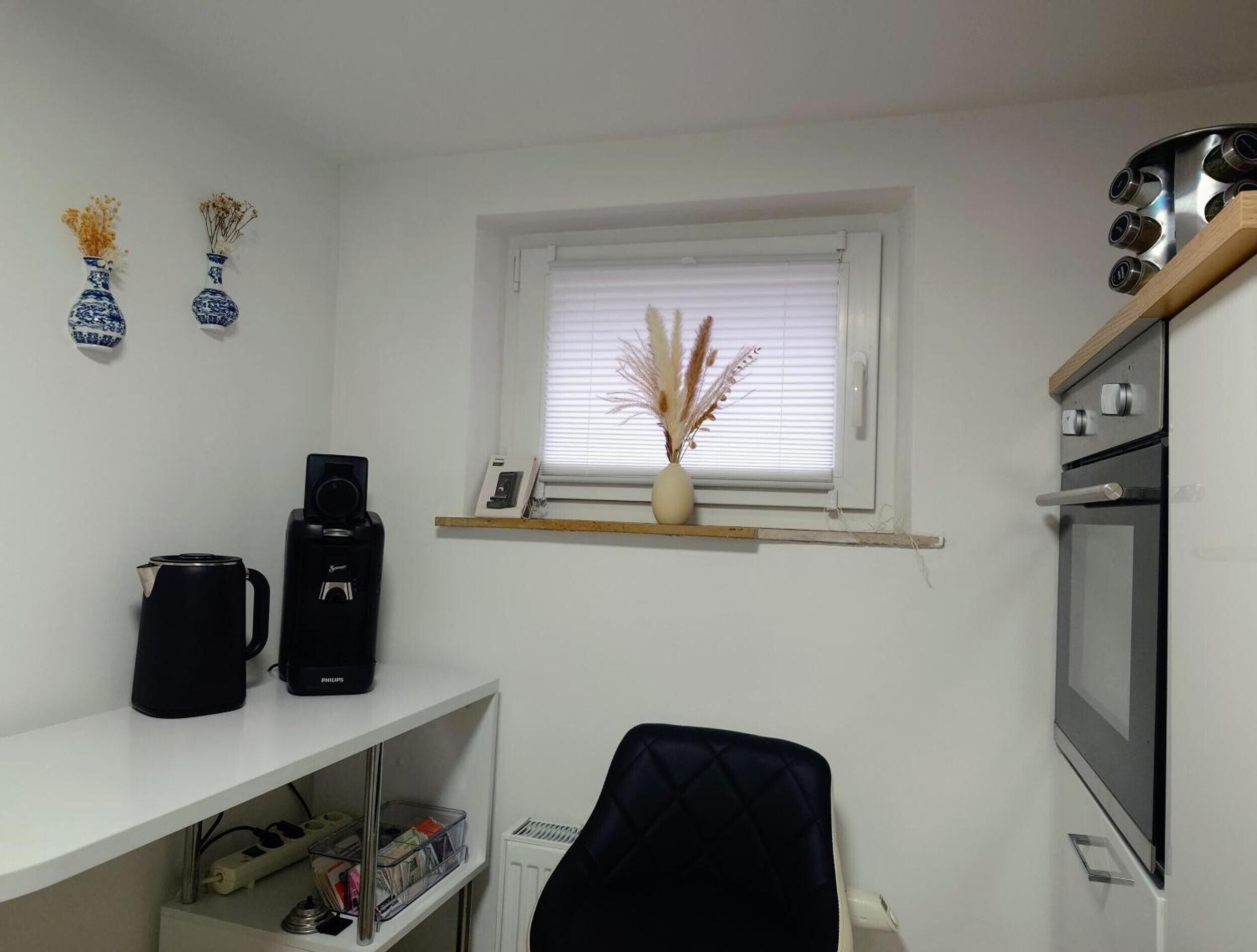 A compact kitchen with a coffee maker, a small shelf with a vase, and a window with blinds.