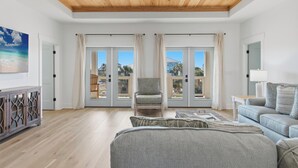 A spacious room with a wooden ceiling, large sliding glass doors leading to a balcony, a comfortable chair, a sofa, and a cabinet. - Beach House - Walking on Water by PHG (St. George Island)