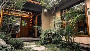 Um edifício em estilo japonês tradicional, com entrada de madeira, caminho de pedra e vegetação exuberante.