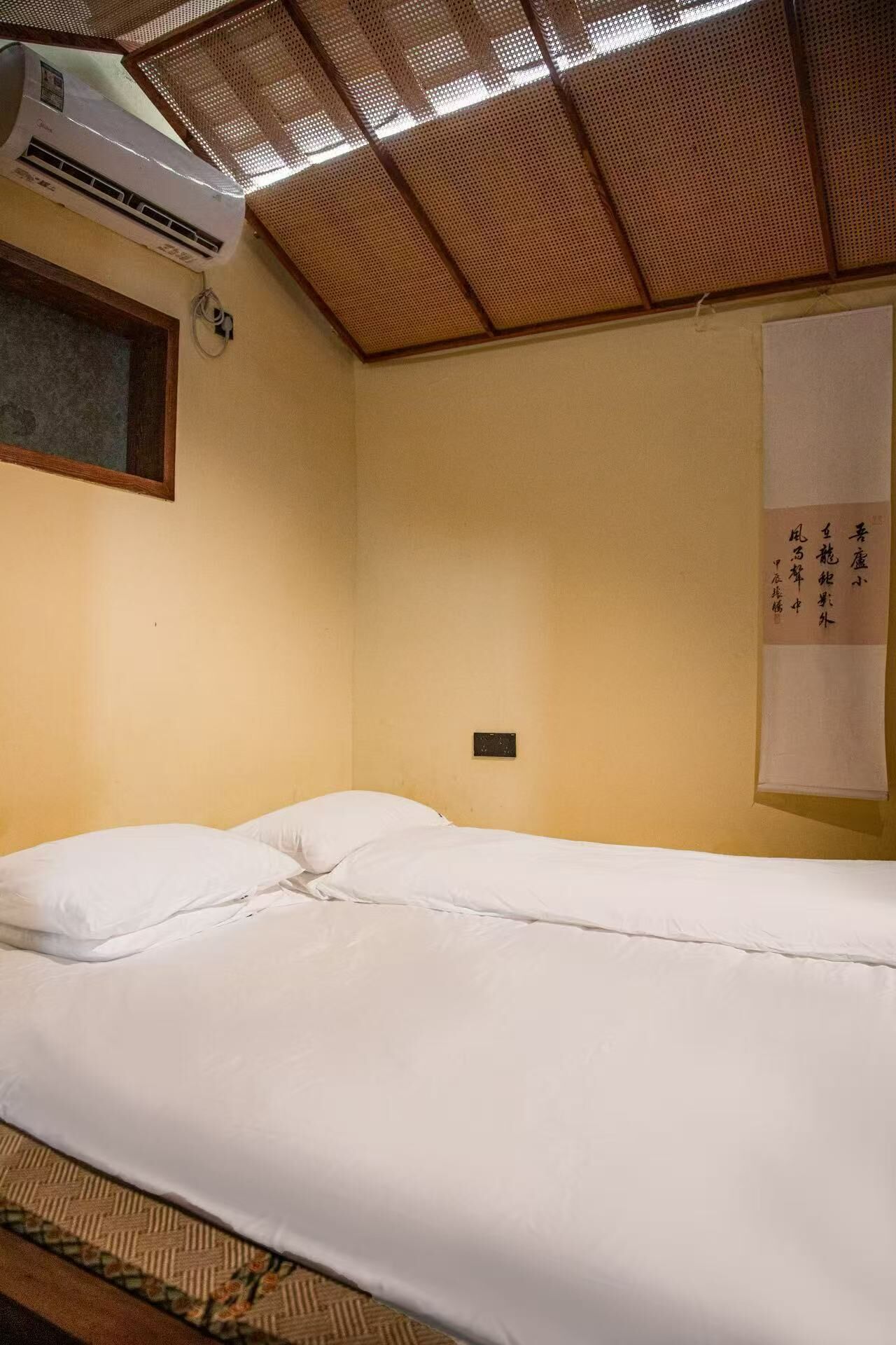 Un lit simple avec une literie blanche, dans une pièce dotée d’un plafond en bois et d’un climatiseur fixé au mur.