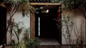 Uma entrada tradicional em estilo japonês, com telhado telhado, porta de madeira e um pequeno jardim com diversas plantas e um caminho de pedra.