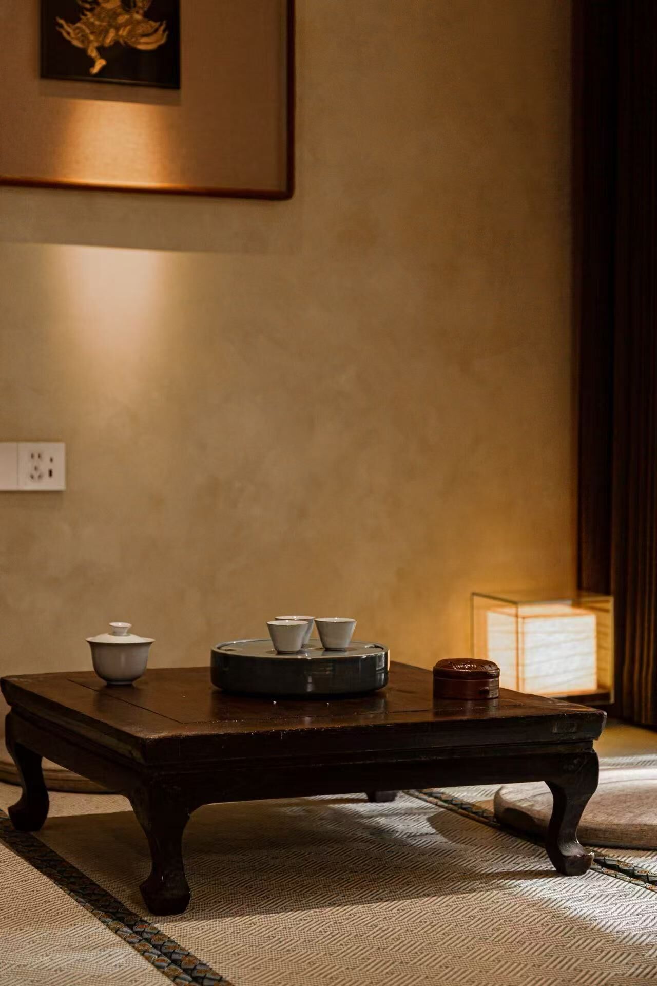 Una habitación tradicional japonesa con suelo de tatami, una mesa de madera oscura con un juego de té y una obra enmarcada en la pared.