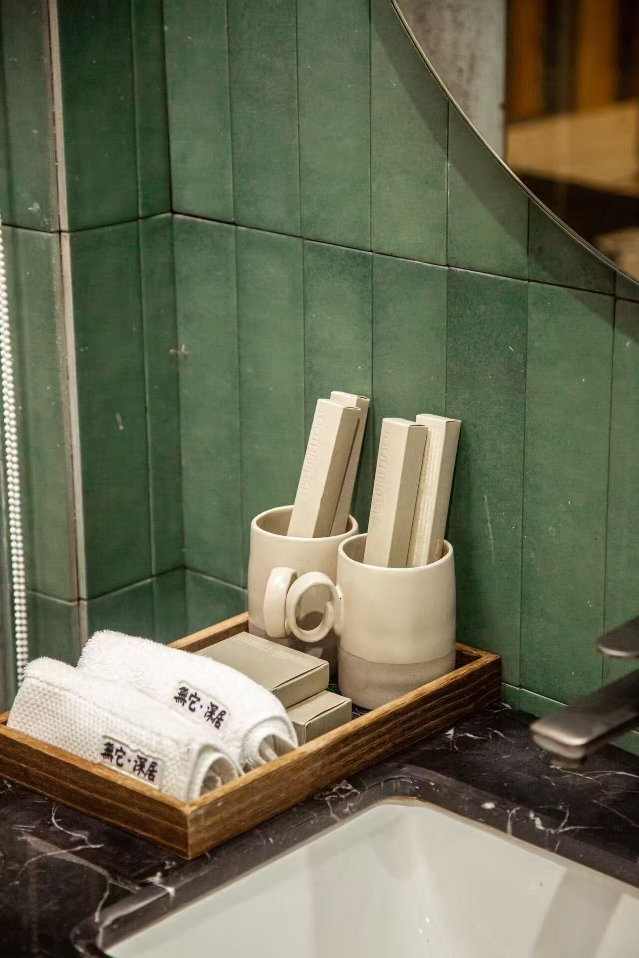 A bathroom with green tiles, a wooden tray holding towels and a toothbrush, and a sink.