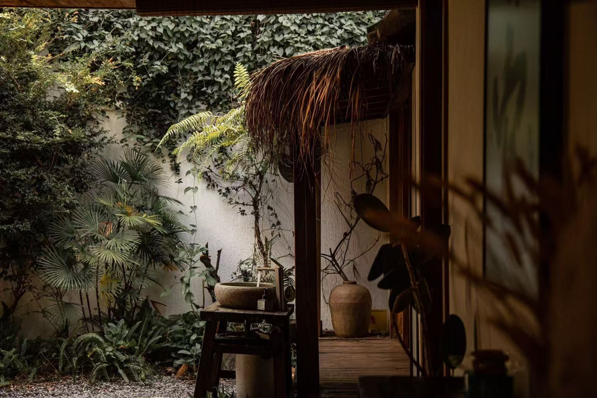 Un jardín japonés tradicional con una estructura de techo de paja, diversas plantas y un cuenco de piedra.