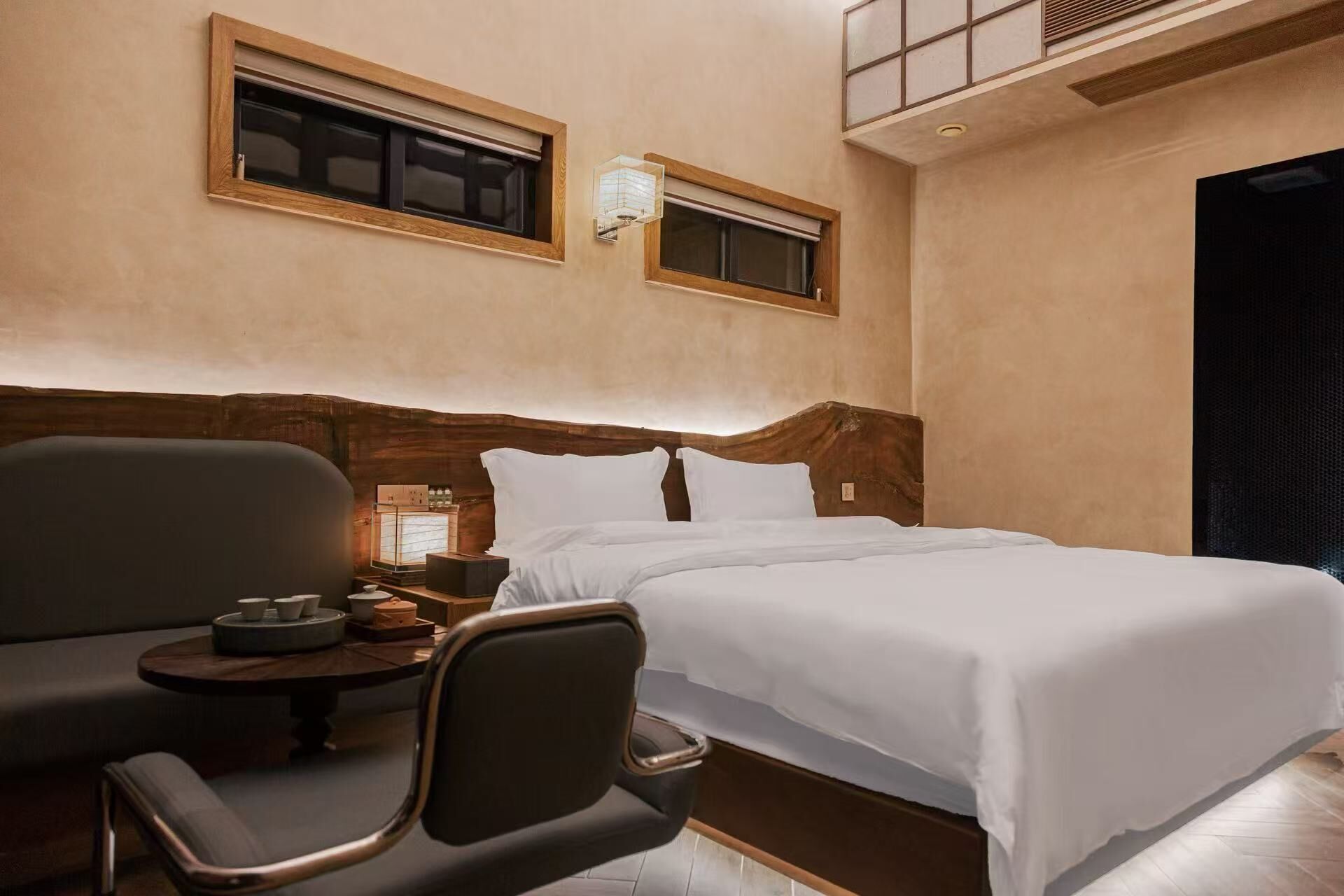 Une chambre d’hôtel avec un lit, une chaise, une petite table et deux fenêtres équipées de stores.