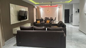 A modern living room with a flat-screen TV, dark sofas, and a dining area with a table and chairs. - Spacious 5-bedroom apartment in serene Haatso Atomic Road with AC (Haatso)