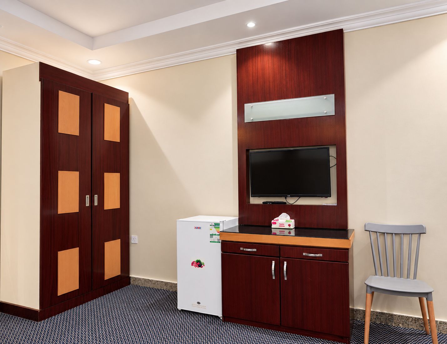 A hotel room with a wardrobe, a small refrigerator, a television mounted on the wall, and a chair.