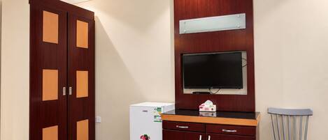 A hotel room with a wardrobe, a small refrigerator, a television mounted on the wall, and a chair.