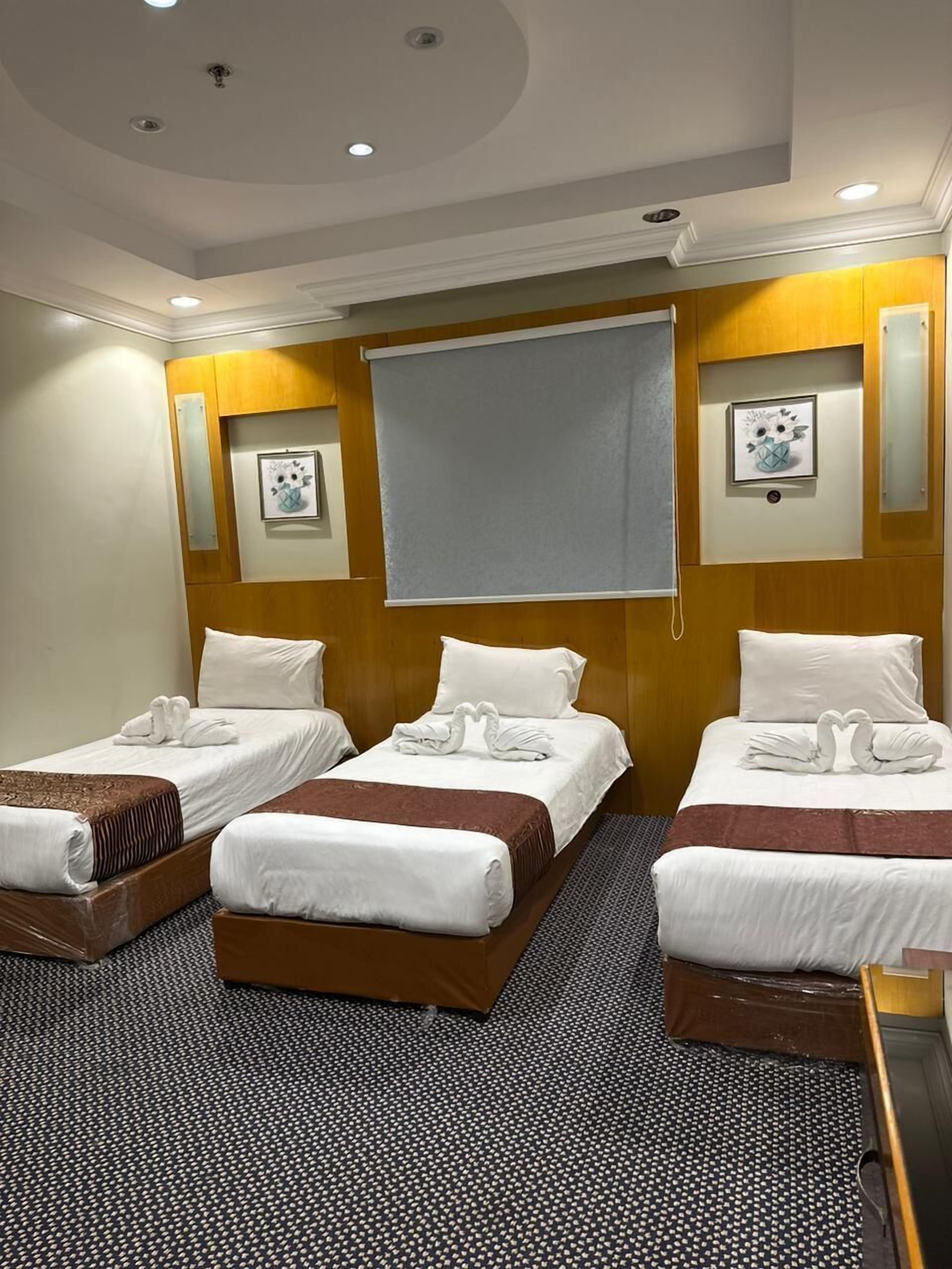 A hotel room with three beds, each with white linens and decorative swan pillows. The room has a wooden headboard, a large window with a blind, and wall-mounted artwork.