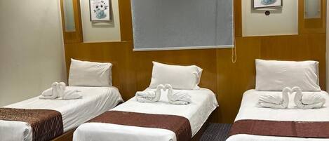 A hotel room with three beds, each with white linens and decorative swan pillows. The room has a wooden headboard, a large window with a blind, and wall-mounted artwork.