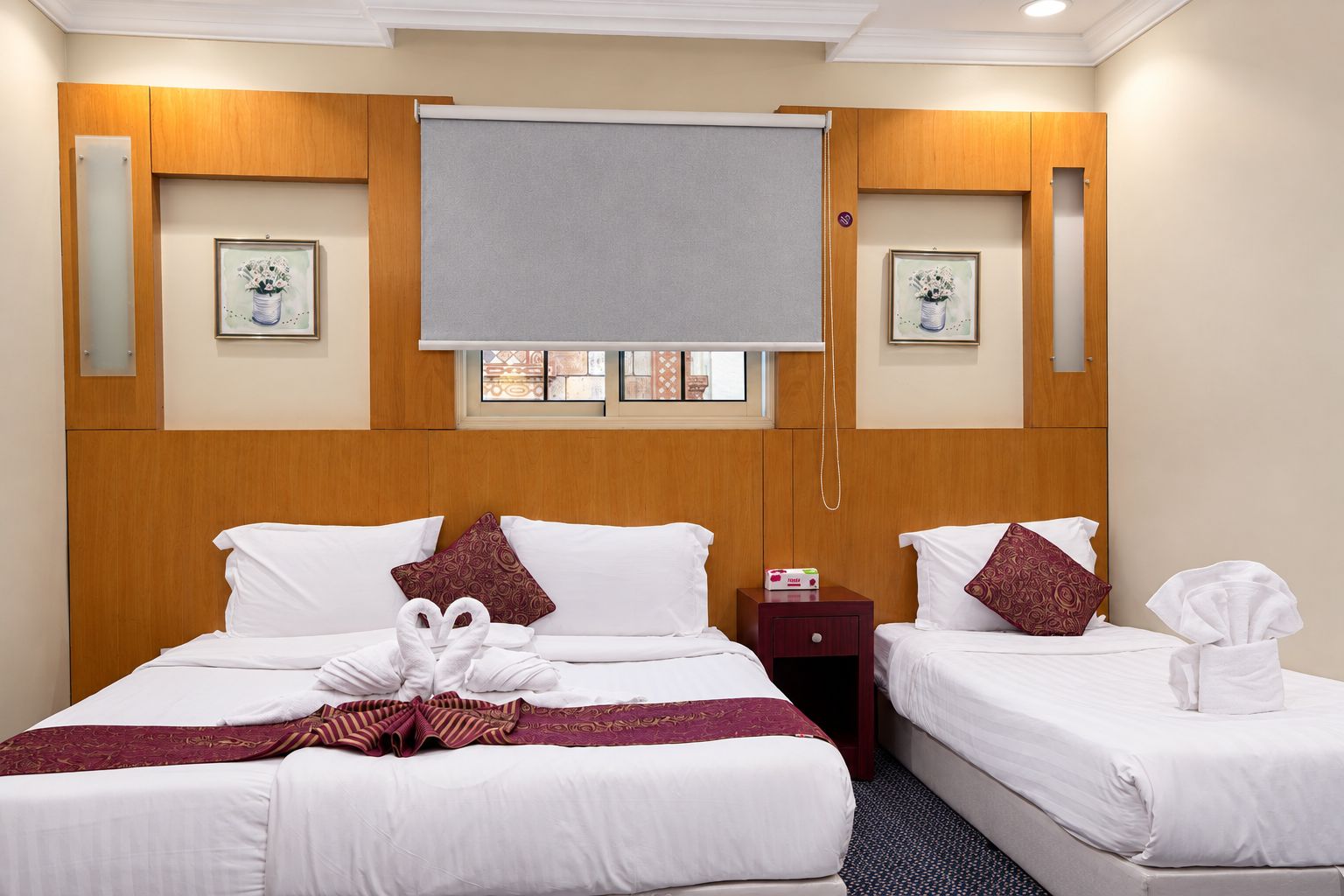 Une chambre d’hôtel avec deux lits, une tête de lit en bois, une fenêtre avec des stores et des tableaux encadrés au mur.