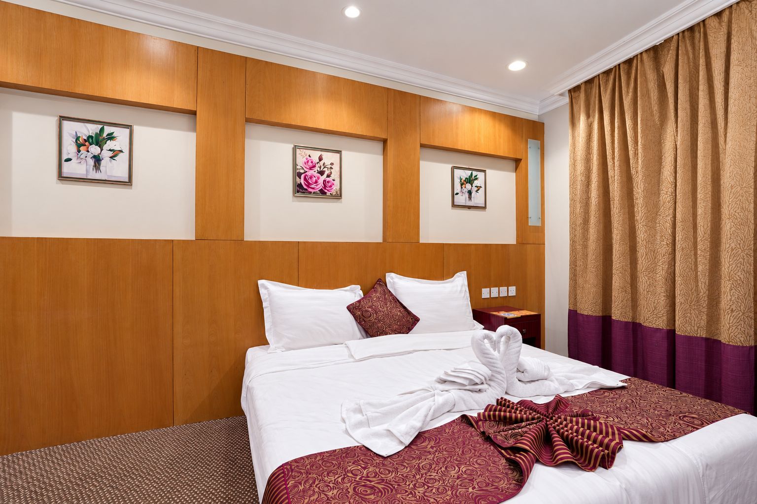 A hotel room with a wooden headboard, a bed with white linens, a decorative pillow, a folded towel, and a patterned throw blanket.