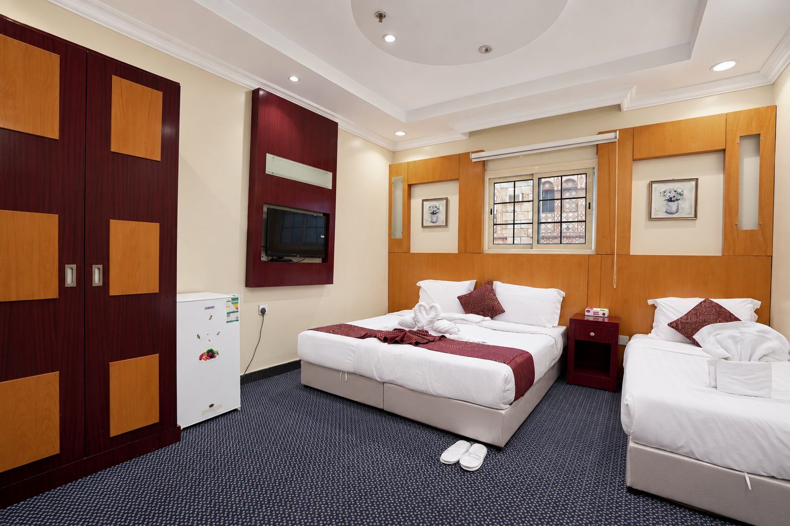 A hotel room with a bed, two bedside tables, a TV, a wardrobe, and a small refrigerator.
