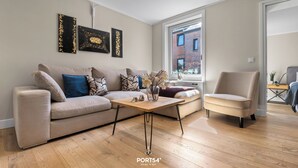 A modern living room with a beige sofa, a coffee table, and a chair. There are three artworks on the wall and a window providing a view of a brick building. - Stadtperle Gold in the heart of Westerland with roof terrace and close to the beach (Sylt)