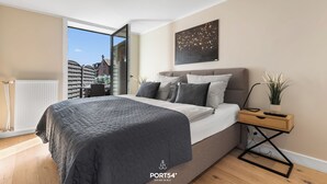 A modern bedroom with a large bed, a nightstand, a potted plant, and a view of buildings through a glass door. - Stadtperle Gold in the heart of Westerland with roof terrace and close to the beach (Sylt)