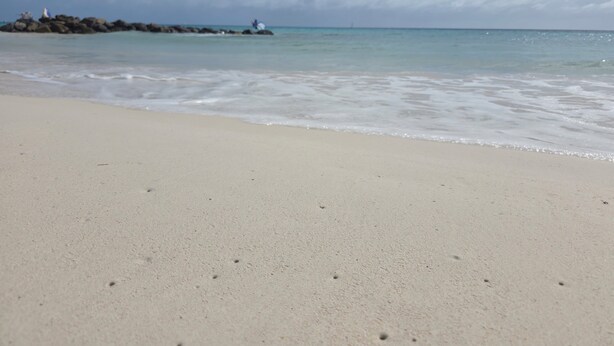 Una playa con un mar tranquilo, un promontorio rocoso y un faro a lo lejos.
