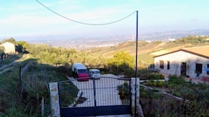 Un chemin de pierres mène à une entrée fermée par un portail, d’où l’on peut admirer un paysage rural et un bâtiment.