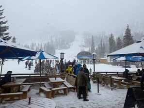 A snowy outdoor area with wooden benches and picnic tables, some covered by blue umbrellas. - Cozy Mountain Condo Steps from the Main Lift (Taos Ski Valley)