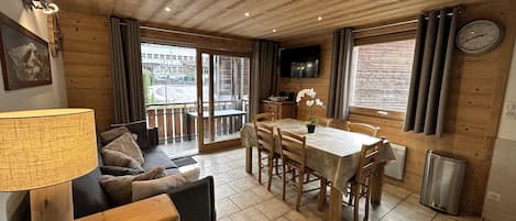 A wooden interior with a sofa, dining table, chairs, a lamp, a clock, and a TV on the wall.
