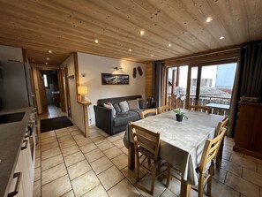 A cozy interior with a dining area, a sofa, and a view through a large window. - NEW RENTAL: KERANGUEN 5 – Village-center apartment, close to slopes and shops – Wi-Fi – Parking (La Clusaz)