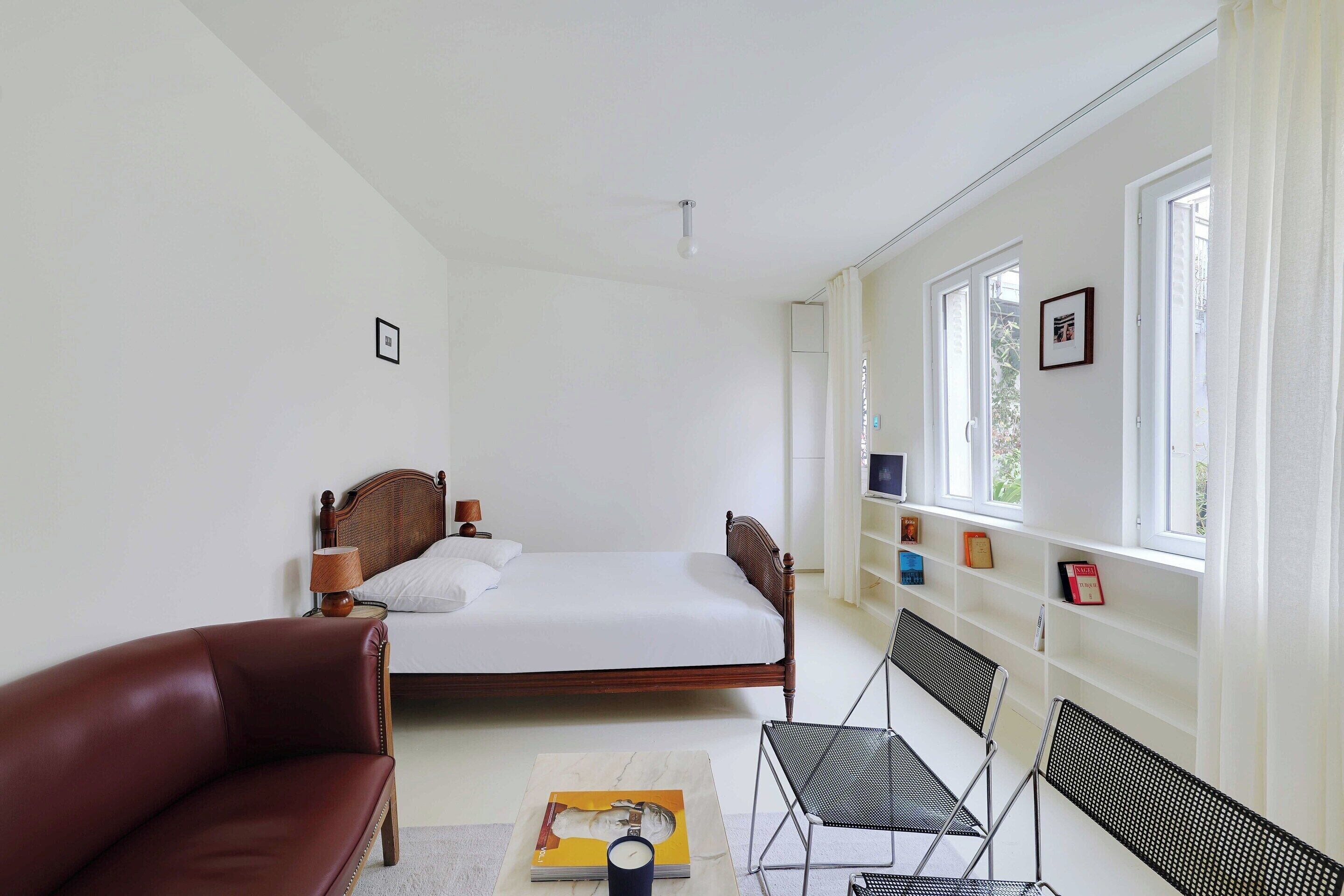 A bedroom with a wooden bed, a red leather chair, a small table with a book, and a window with curtains.