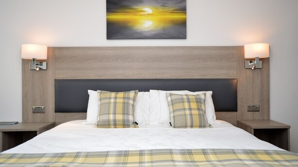 A hotel room with a large bed, two bedside tables, a headboard, and a wall-mounted painting of a sunset.