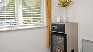 A room with a minibar, a refrigerator, a window with blinds, and a vase with flowers on a shelf.