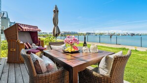 Eine Holzterrasse mit Korbmöbeln, einem Tisch mit Obstschale und Blick auf ein Gewässer.