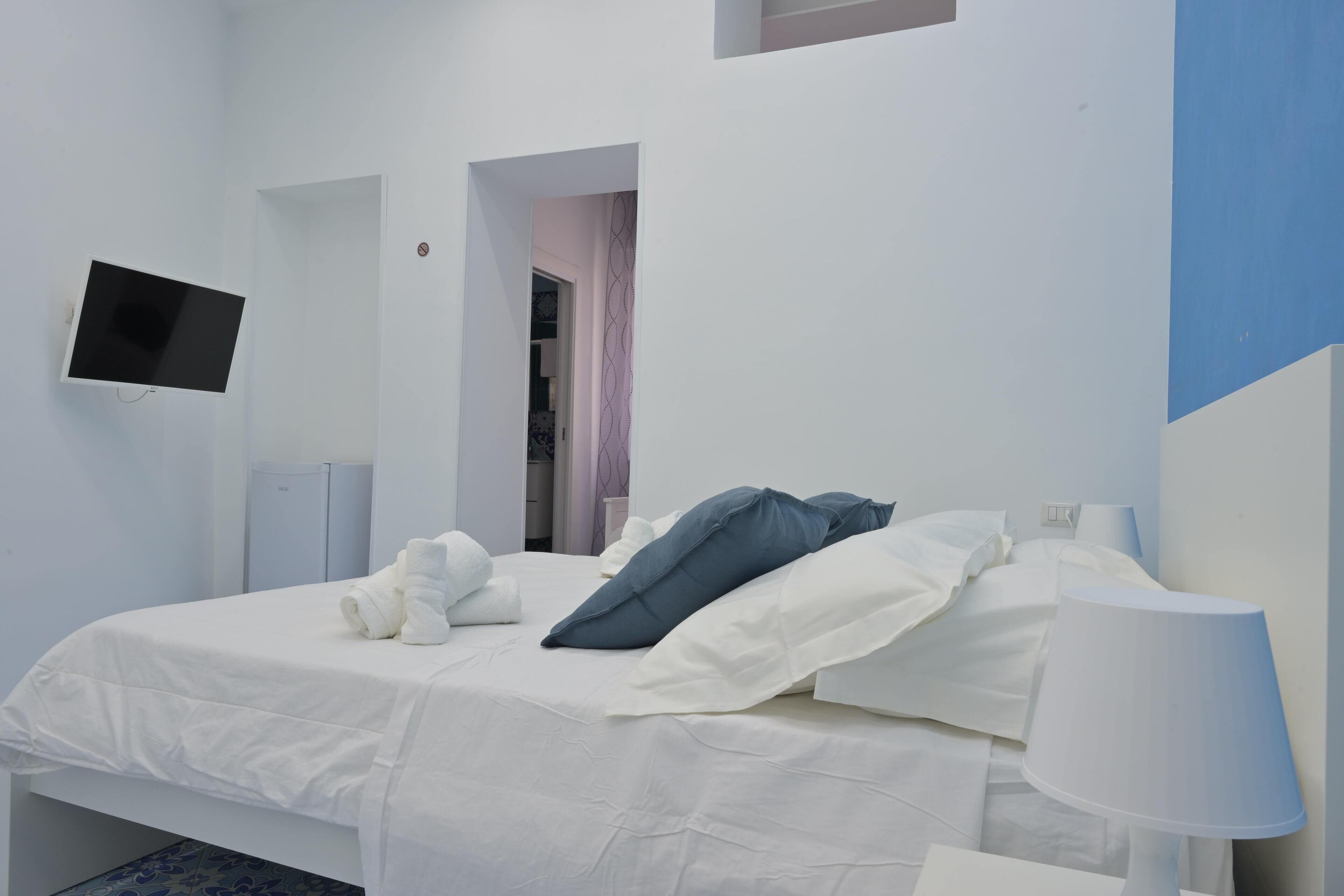 A neatly made bed with white bedding and a single blue pillow, a bedside lamp, and a flat-screen TV mounted on the wall.