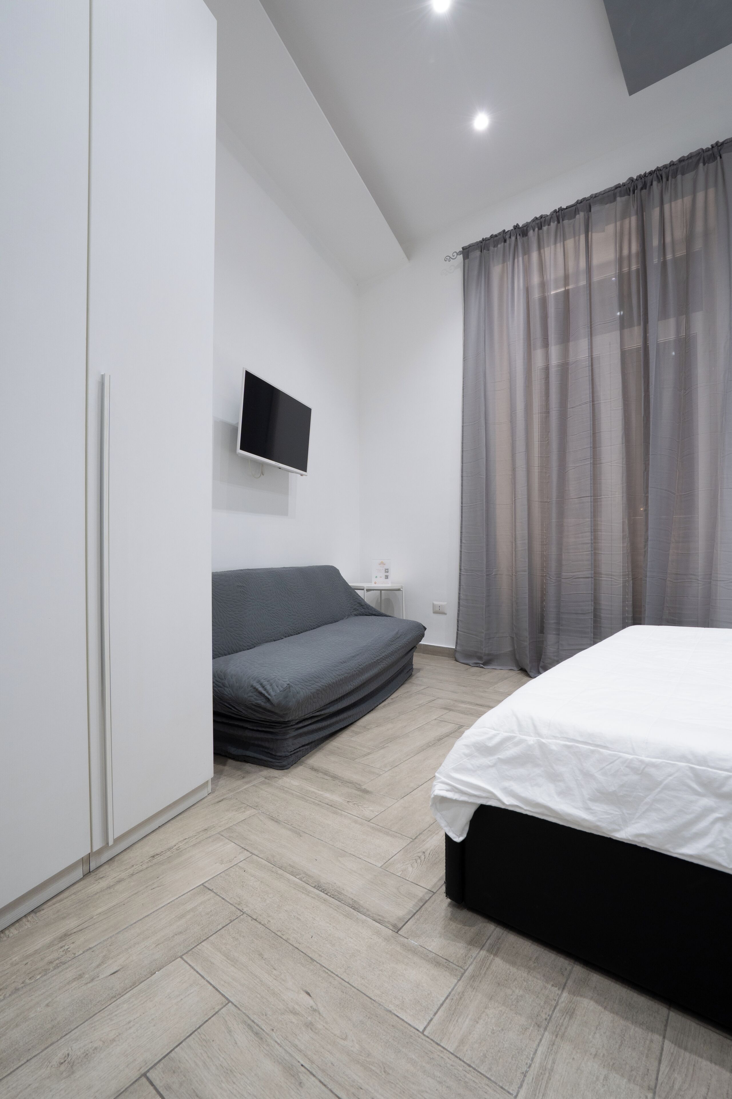 A modern bedroom with a bed, a sofa, a TV, and a window with sheer curtains.