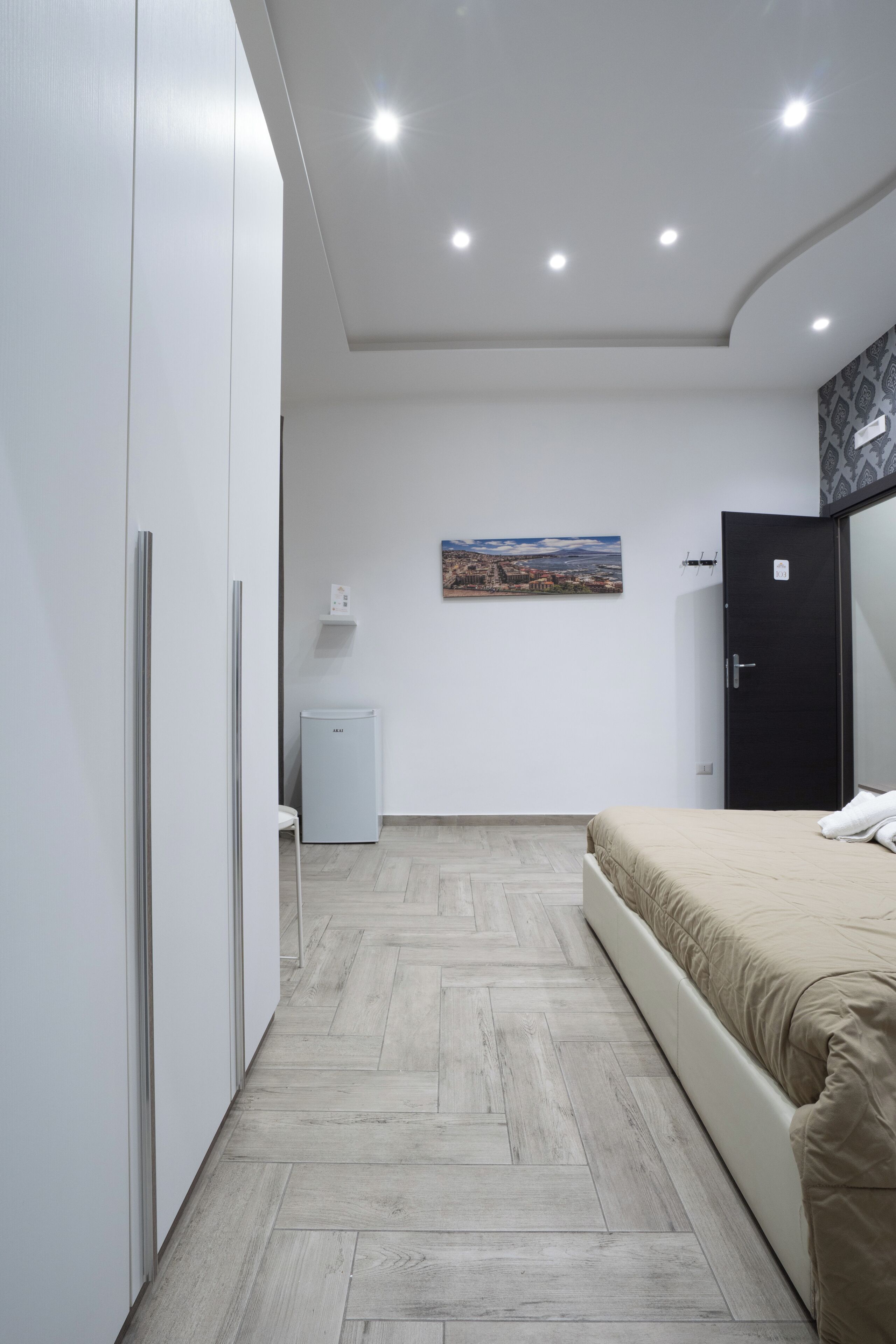 A modern bedroom with a bed, a wardrobe, a small white cabinet, and a painting on the wall.