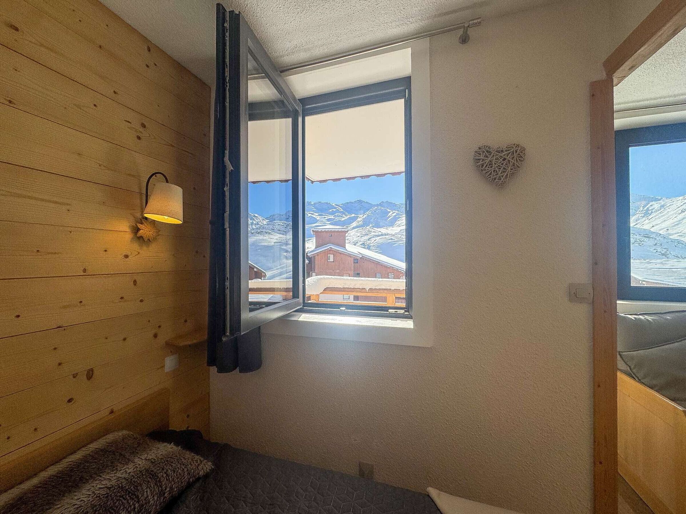 Quarto com paredes de madeira, janela com vista para montanhas cobertas de neve, luminária fixada na parede e um enfeite em formato de coração.