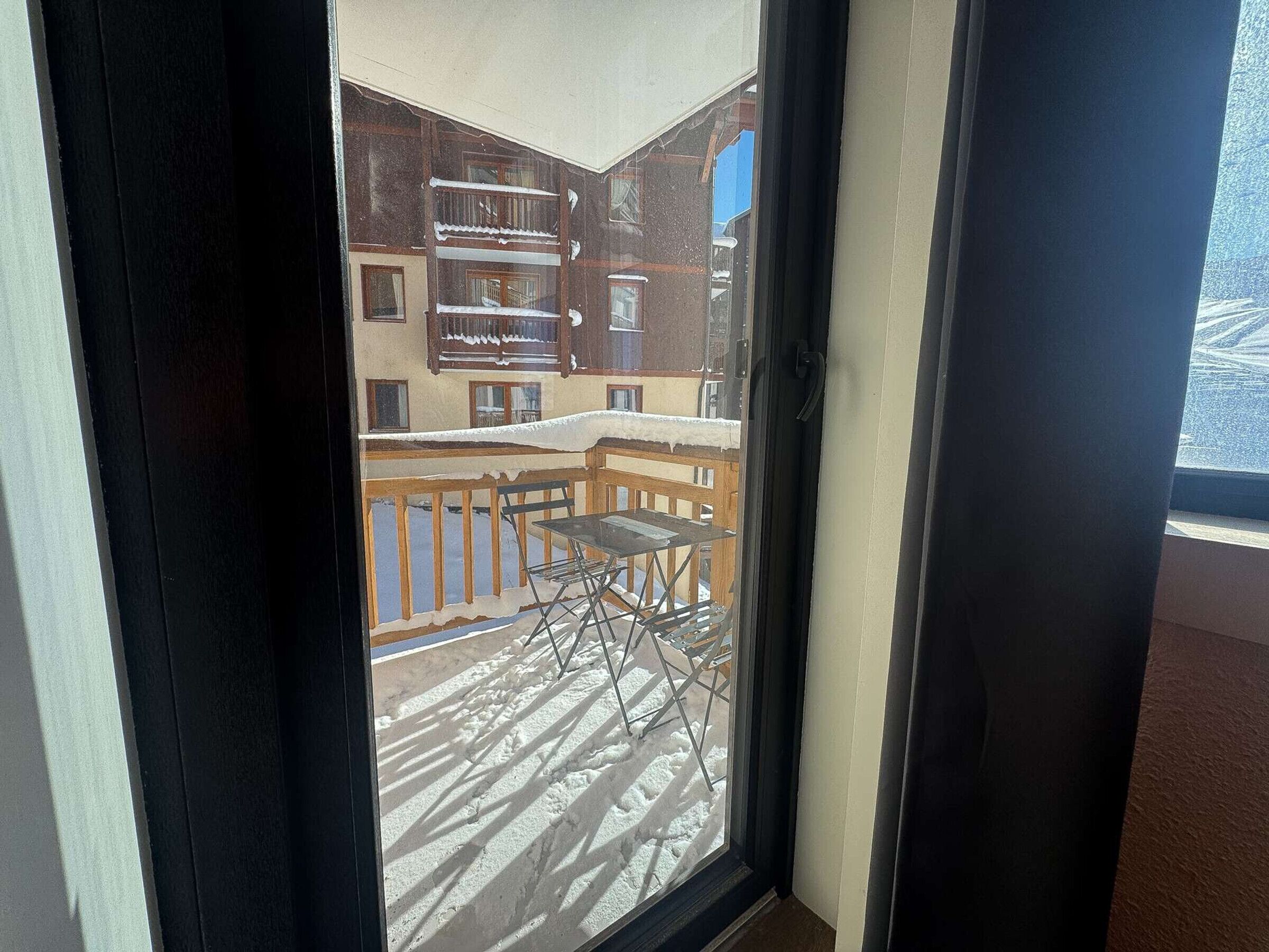 Blick durch ein Fenster auf einen Balkon mit Tisch und Stühlen, verschneite Erde und ein Gebäude mit zahlreichen Fenstern und Balkonen.