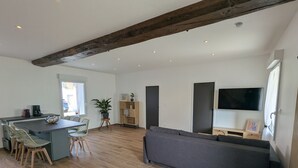 Een moderne woonkamer met houten vloer, een grijze bank, een zithoek met stoelen, een kleine keuken en een tv aan de muur.