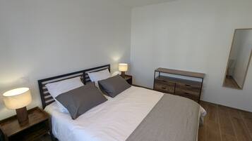 Een netjes opgemaakt bed met een witte dekbedovertrek en twee kussens, een houten nachtkastje met een lamp, een houten dressoir en een spiegel aan de muur.