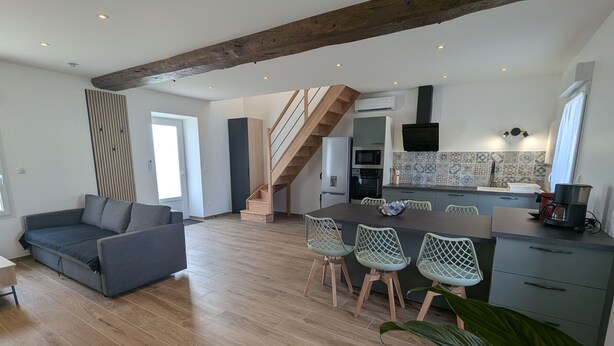 Een moderne woonruimte met een houten vloer, een grijze bank, een eethoek met stoelen, een keuken met een betegelde spatwand, en een trap.