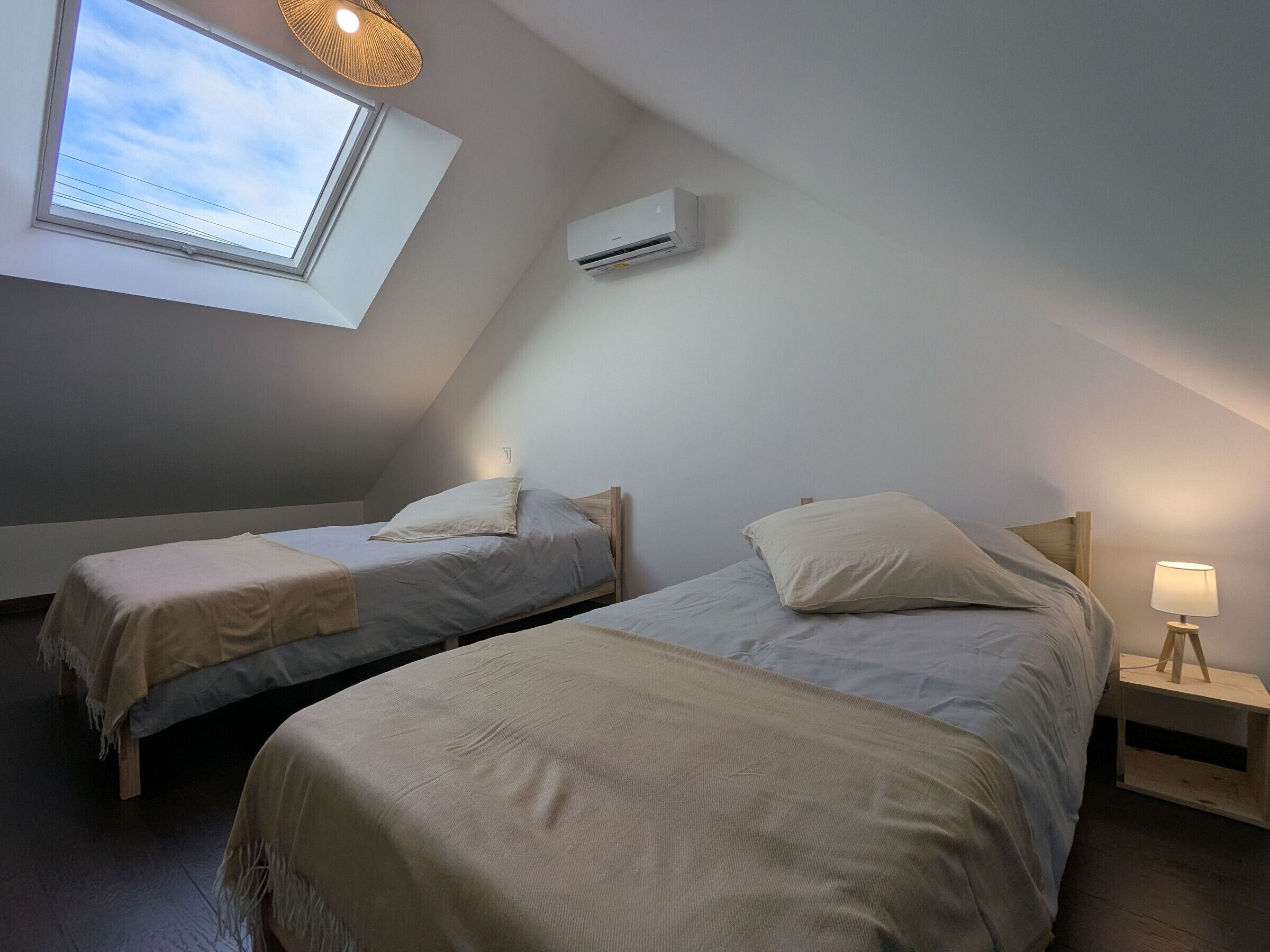 Une petite chambre confortable avec deux lits, une lucarne, un climatiseur et une lampe de chevet.