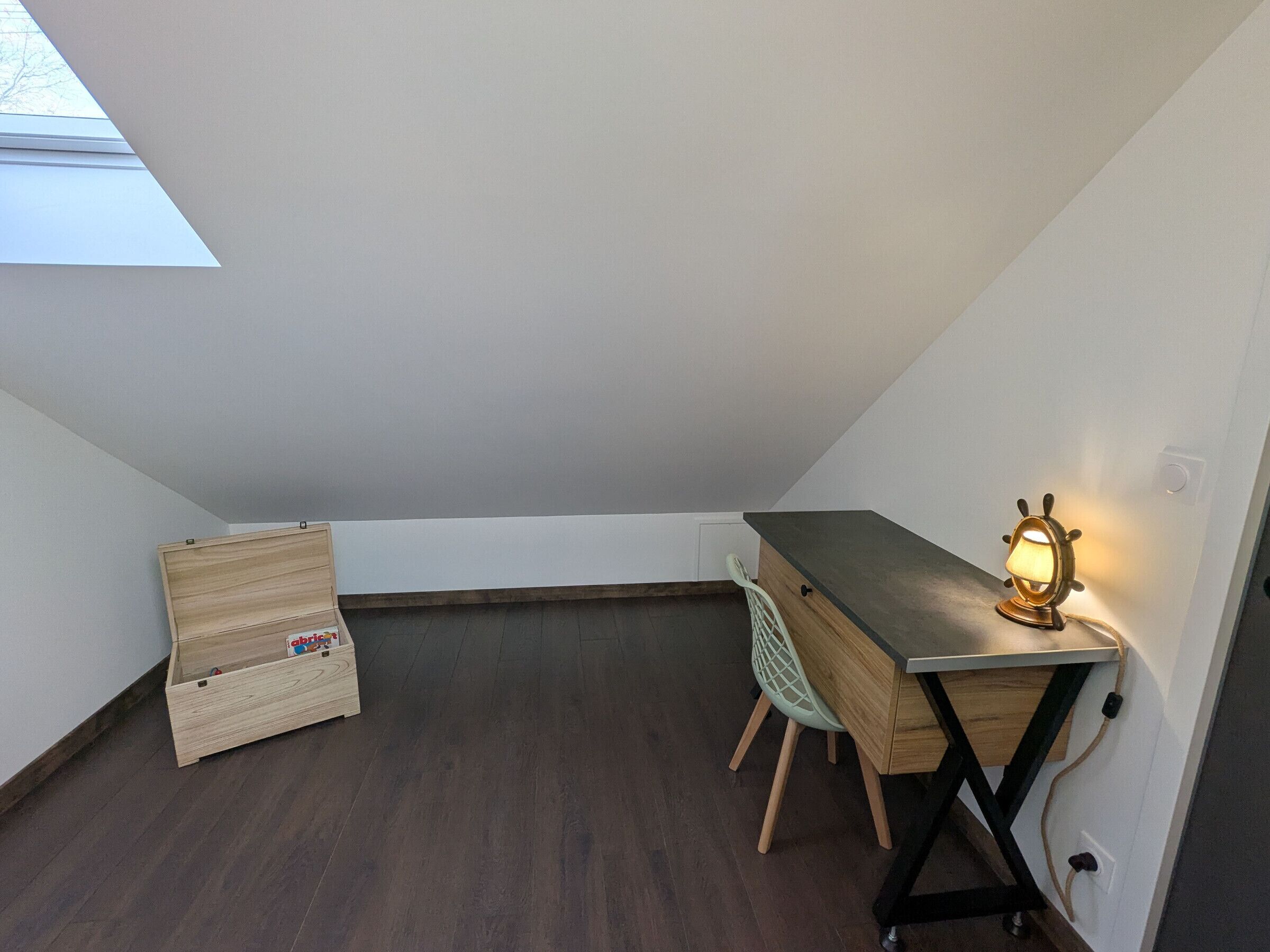 A small, dimly lit room with a slanted ceiling, a wooden desk, a chair, and a small lamp.